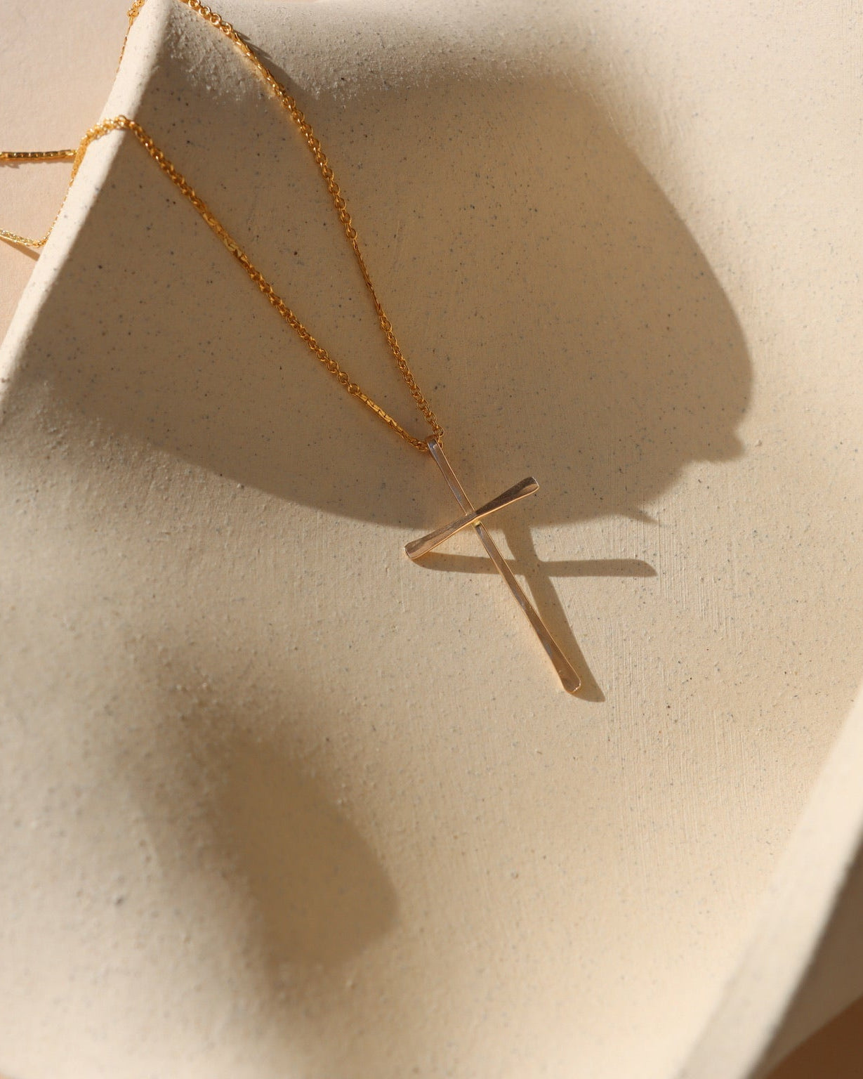 Grace Cross Necklace in 14k gold fill featuring a modern cross pendant on a delicate chain displayed on a textured ceramic surface.