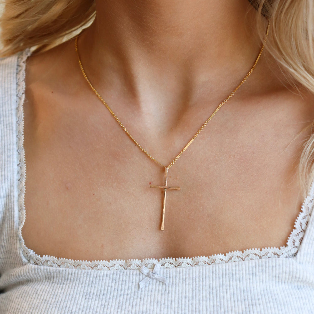 Model is wearing a 14k gold fill thin bar and link chain featuring a 1 1/2" hammered cross pendant. Model is wearing a gray low neck top. 