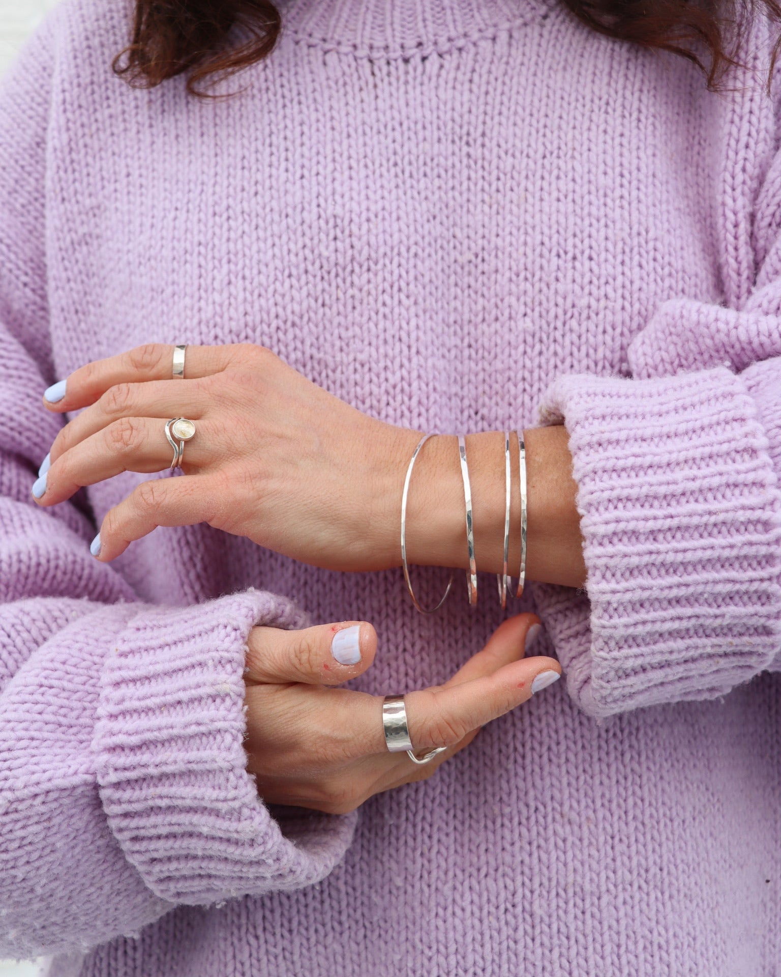 A close-up of sterling silver bangles stacked on a wrist, paired with silver statement rings against a cozy lavender knit turtleneck sweater.