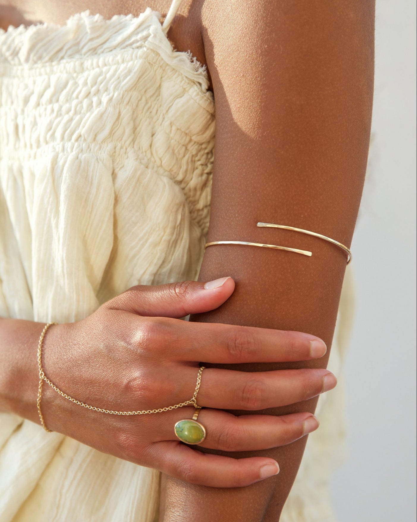 Close-up shot of a woman in a white textured dress resting her hand on her arm, styled with boho gold jewelry: a sleek open wrap upper arm cuff, a delicate gold hand chain connecting her wrist to a middle-finger ring, and an oval green gemstone ring on her index finger