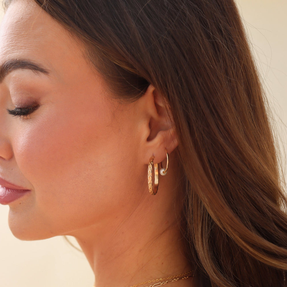 Close-up of a woman wearing gold hoop earrings and a necklace against a neutral background