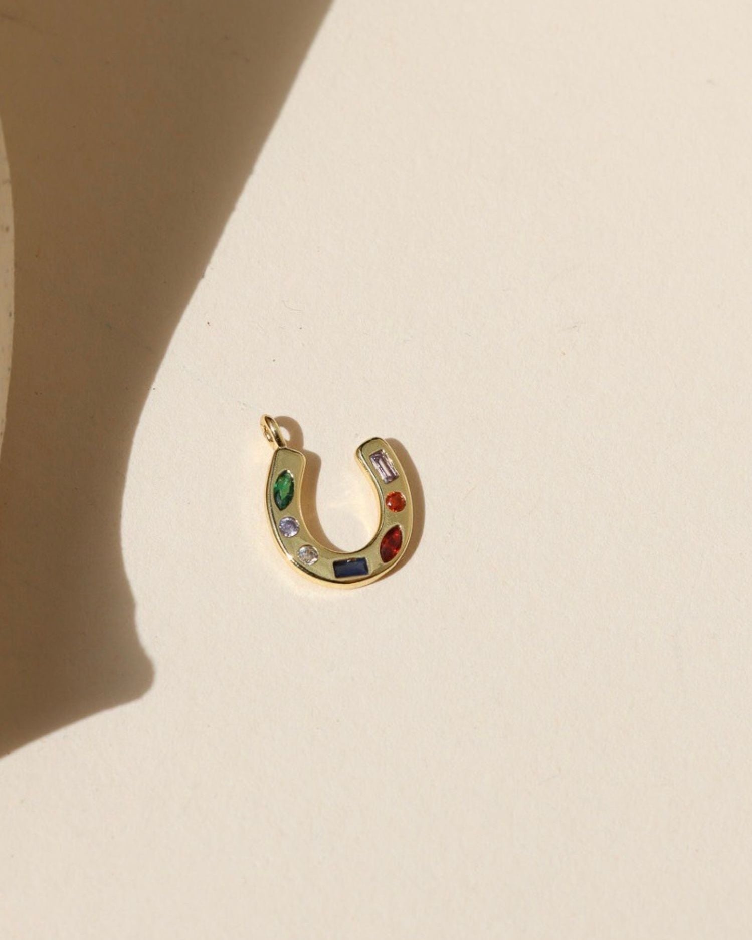 Gold horseshoe shaped charm with green, clear, blue, and red CZ gems embedded within, laid on a beige background in the natural sunlight. 