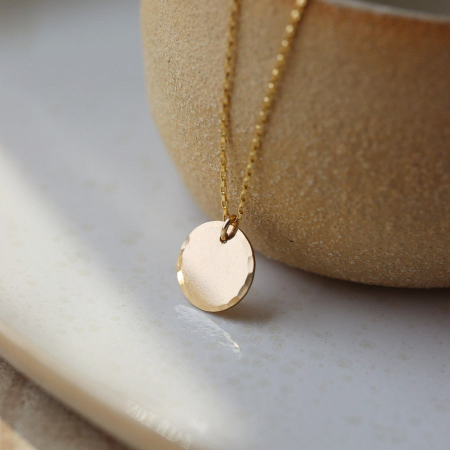 14k gold fill Mini coin necklace placed on a tan bowl in the sunlight. This Necklace features a coin disc hammered on the outside of the circle. it slides around on the coin necklace chain.