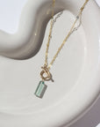 14k gold fill Flynn Mini Toggle Necklace laid on a tan plate in the sunlight. This Necklace Features a Simple Toggle connector with a Dangle Fluorite tube gemstone . The toggle is held together with the Sylvie chain.