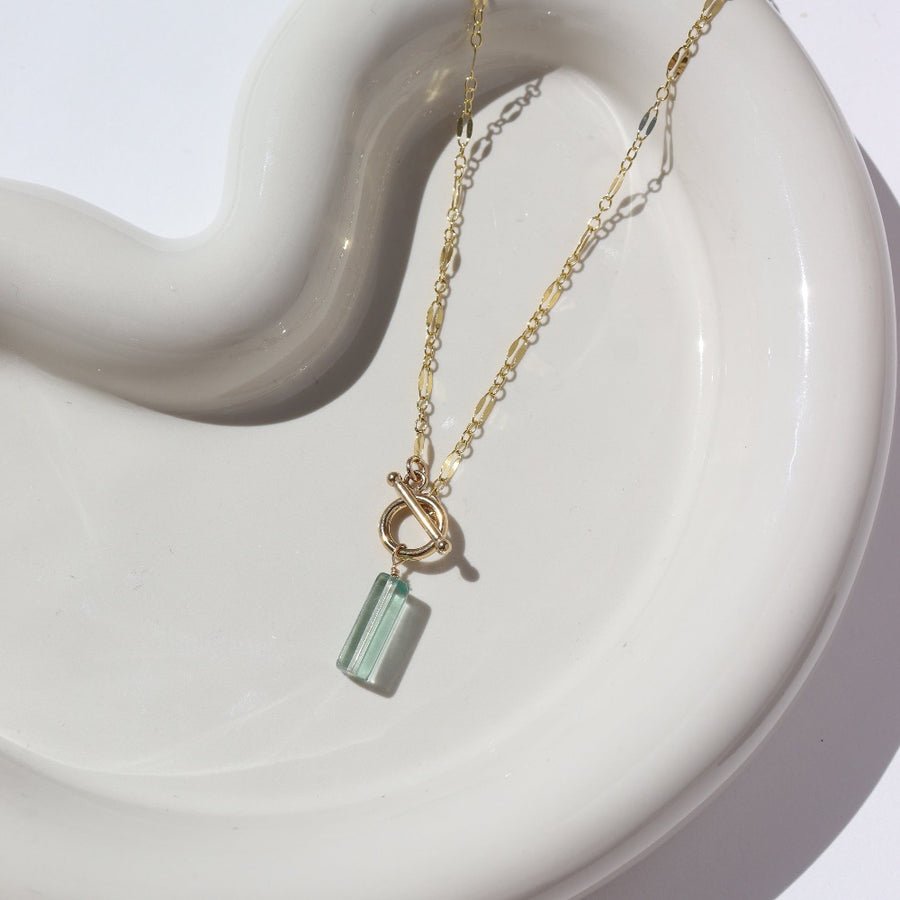 14k gold fill Flynn Mini Toggle Necklace laid on a tan plate in the sunlight. This Necklace Features a Simple Toggle connector with a Dangle Fluorite tube gemstone . The toggle is held together with the Sylvie chain.
