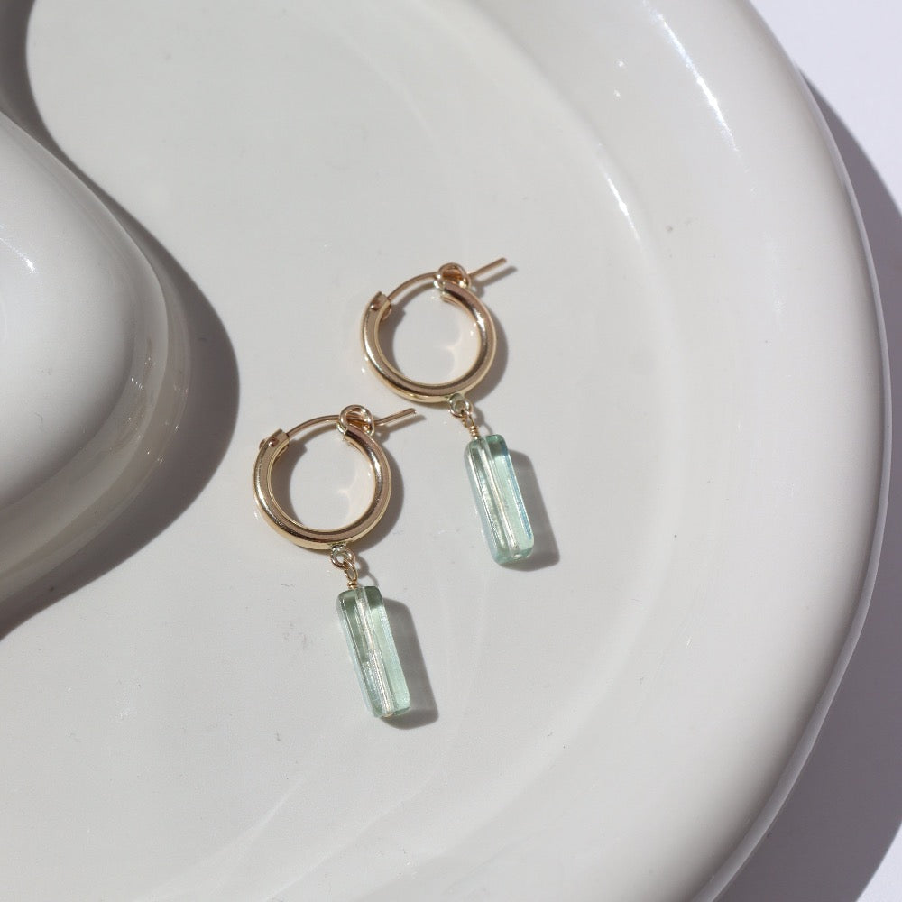 14k gold fill Flynn Hoops laid on a tan plate in the sunlight. These earrings feature a hoop like earring with a dangle of a Fluorite tube gemstone.