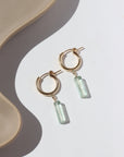14k gold fill Flynn Hoops laid on a tan plate in the sunlight. These earrings feature a hoop like earring with a dangle of a Fluorite tube gemstone.