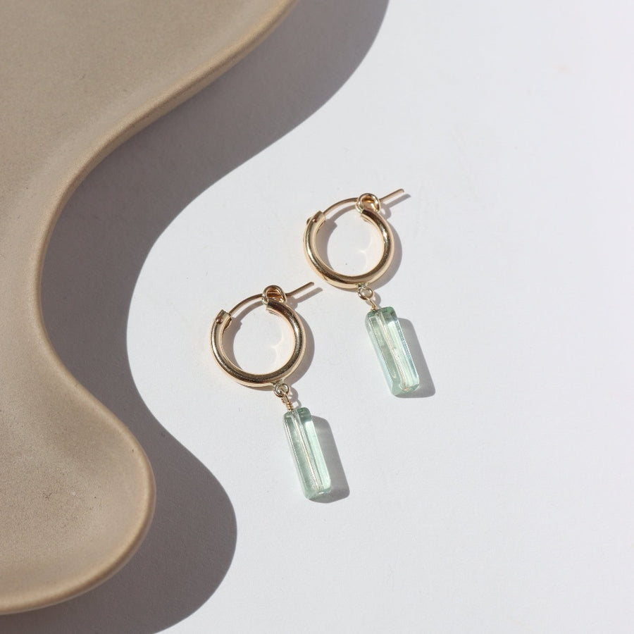 14k gold fill Flynn Hoops laid on a tan plate in the sunlight. These earrings feature a hoop like earring with a dangle of a Fluorite tube gemstone.