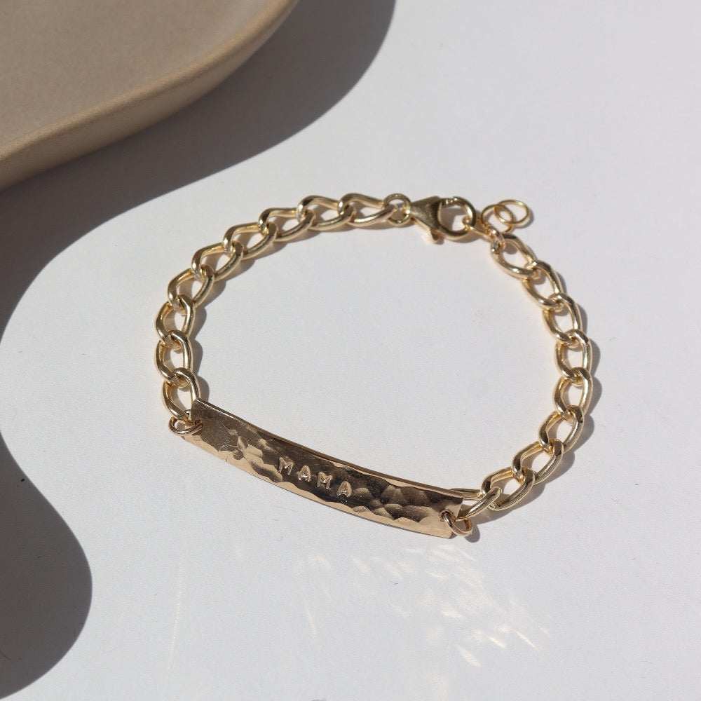 14k gold fill Alexandra Name Plate laid on a tan plate in the sunlight. This Bracelet features a simple hammered Name plate that you get to customize with any character of your choice with 8 initials. The plate is connected by the Alexandra chain.