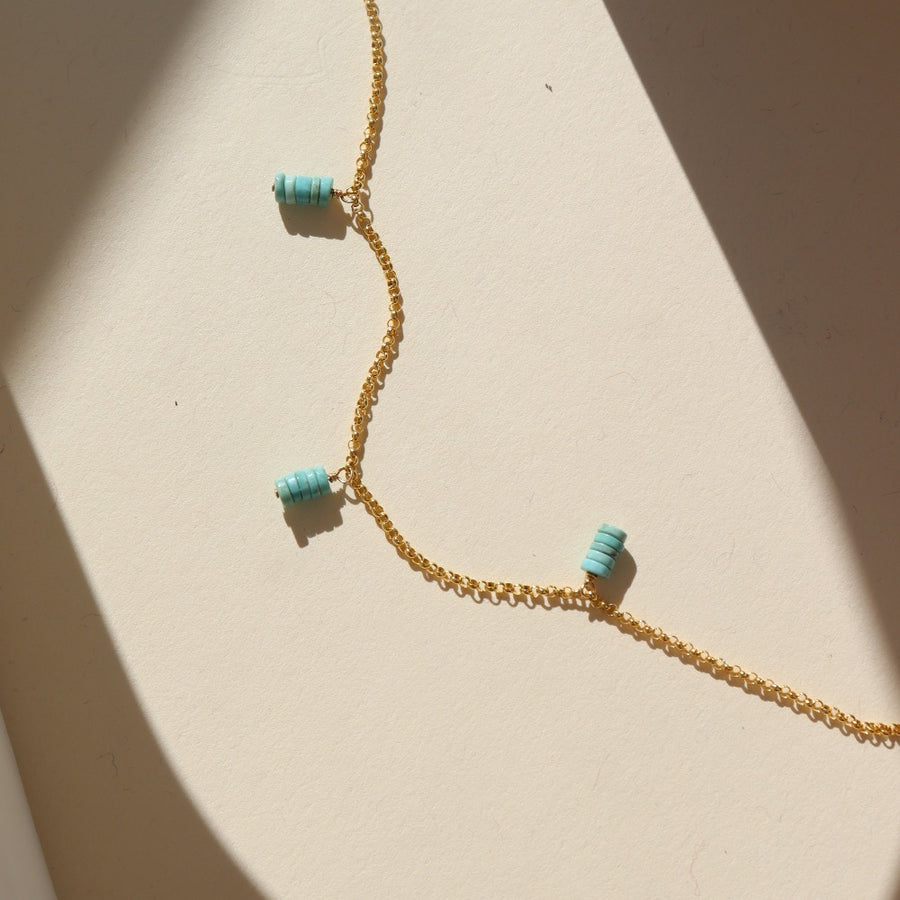A simple 14k gold fill chain adorned with three charms of stacked turquoise stones, laid out on a cream colored backdrop