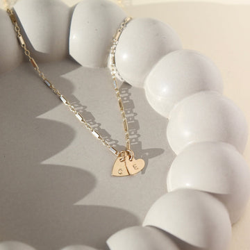 two dainty heart charms with initials stamped on them, on a necklace chain, photographed on a ceramic dish