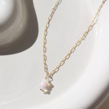 14k gold fill Gender Reveal Necklace laid on a white plate in the sunlight. This necklace features a beautiful mini paper clip chain that is met together with either a pink or blue opal gemstones. 