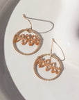 14k gold fill Gaia Earrings laid on a white plate in the sunlight. This earrings features a hook like earring with a spiral hoop featuring pink zircon gemstones.