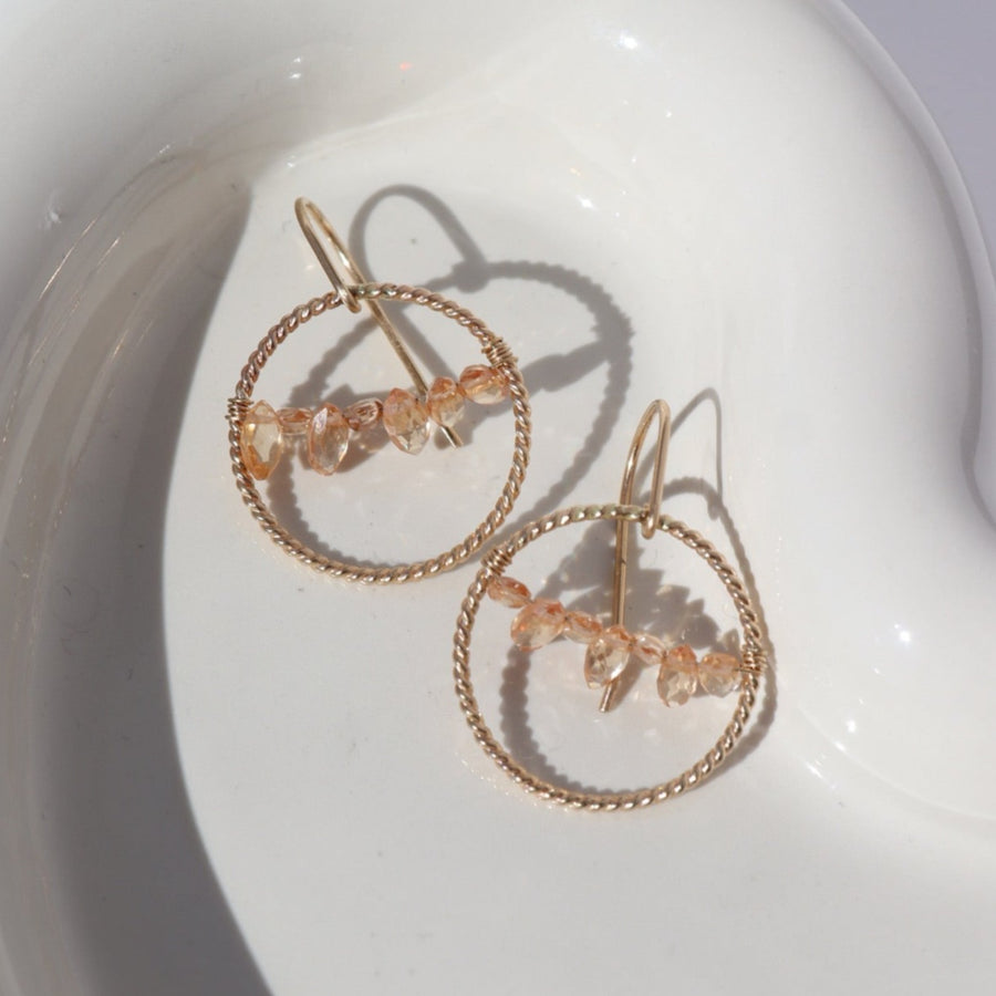14k gold fill Gaia Earrings laid on a white plate in the sunlight. This earrings features a hook like earring with a spiral hoop featuring pink zircon gemstones.