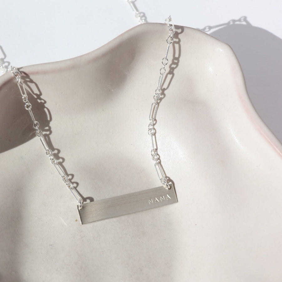 925 sterling silver Matriarch Necklace laid on a tan plate in the sunlight. This necklace features either the simple chain and or the April chain and the word "NANA". you can have the option of a smoother or hammered.