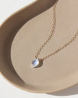 faceted marquise moonstone gemstone anchored on a solid gold chain and laying in the sunlight on a ceramic dish.