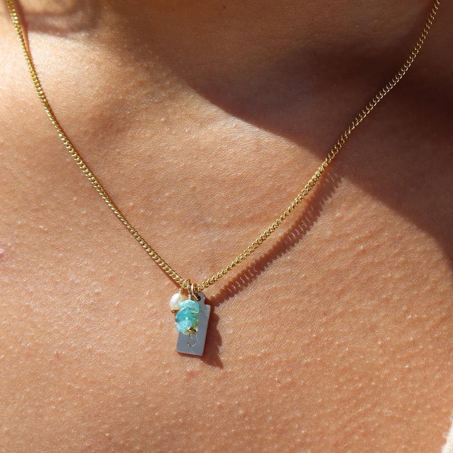 14k gold fill chain necklace featuring an initial stamped tag, three blue opal chips, and a  pearl charm, photographed on a model wearing a cream vest