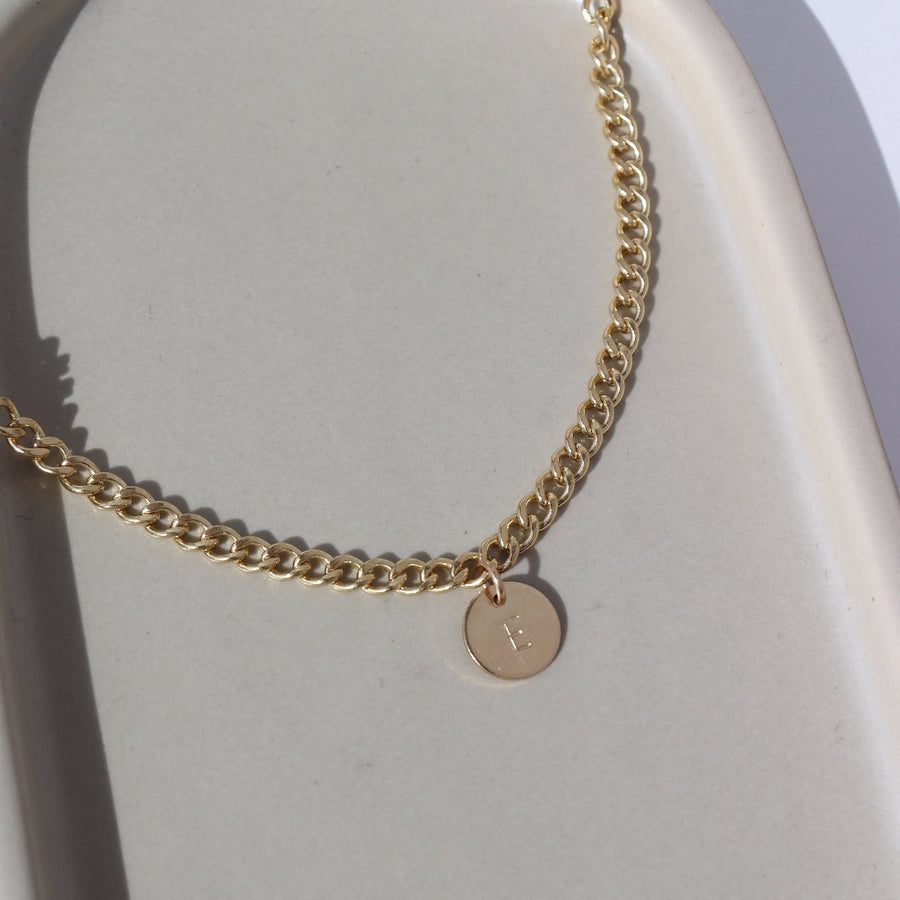 14k gold fill Modern Muse Initial Necklace laid on a tan plate in the sunlight. This Necklace feature our Demi Alexandra chain featuring a charm initial of your choice.