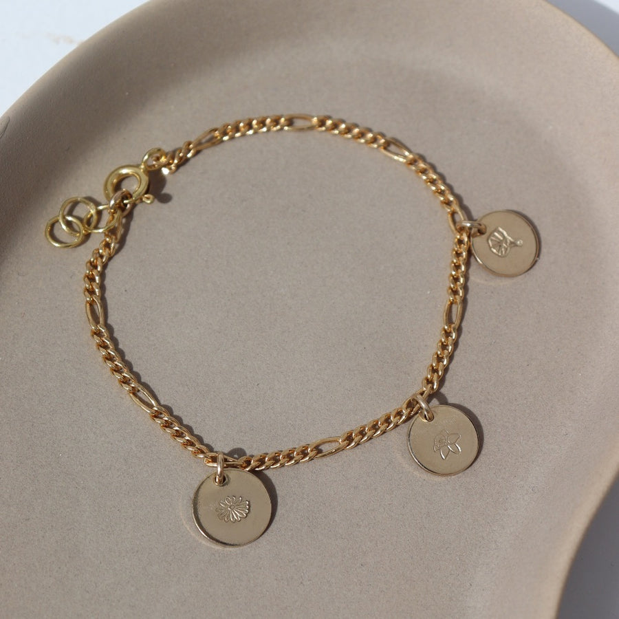 14k gold fill Birth Flower Charm Bracelet laid on a tan plate in the sunlight. This Bracelet feature the Demi Alexandra chain with as many birth flower charms as you want.