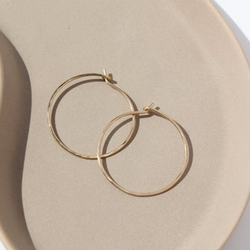 14k solid gold Organic Hoops by Token Jewelry. The hoops are a circle shape with hammered solid gold wire, photographed on a tan ceramic dish