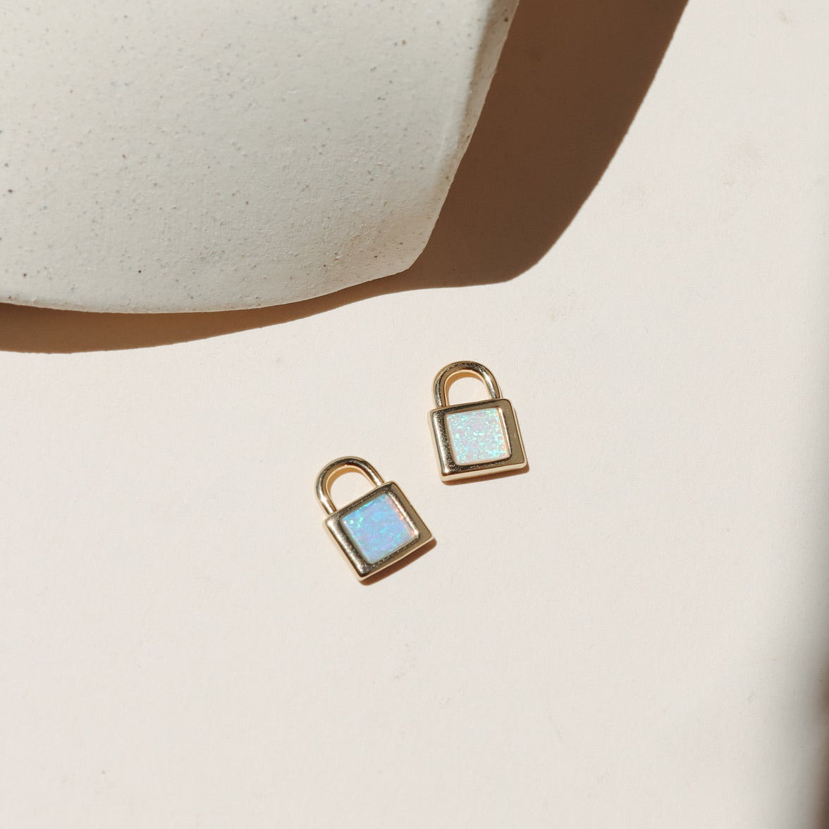 Gold padlock charm with shimmering white and blue opal stones on a neutral surface in warm natural light.