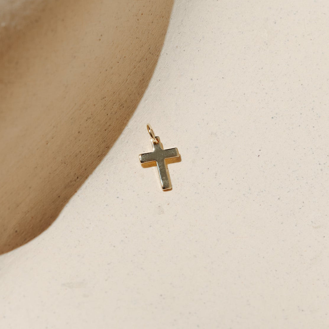 Small smooth gold cross charm displayed on a neutral surface in warm natural light.