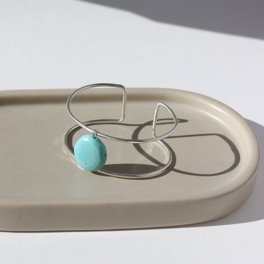 sterling silver double wire cuff bracelet featuring a large turquoise stone wired to the middle. Photographed on a taupe dish in the sunlight