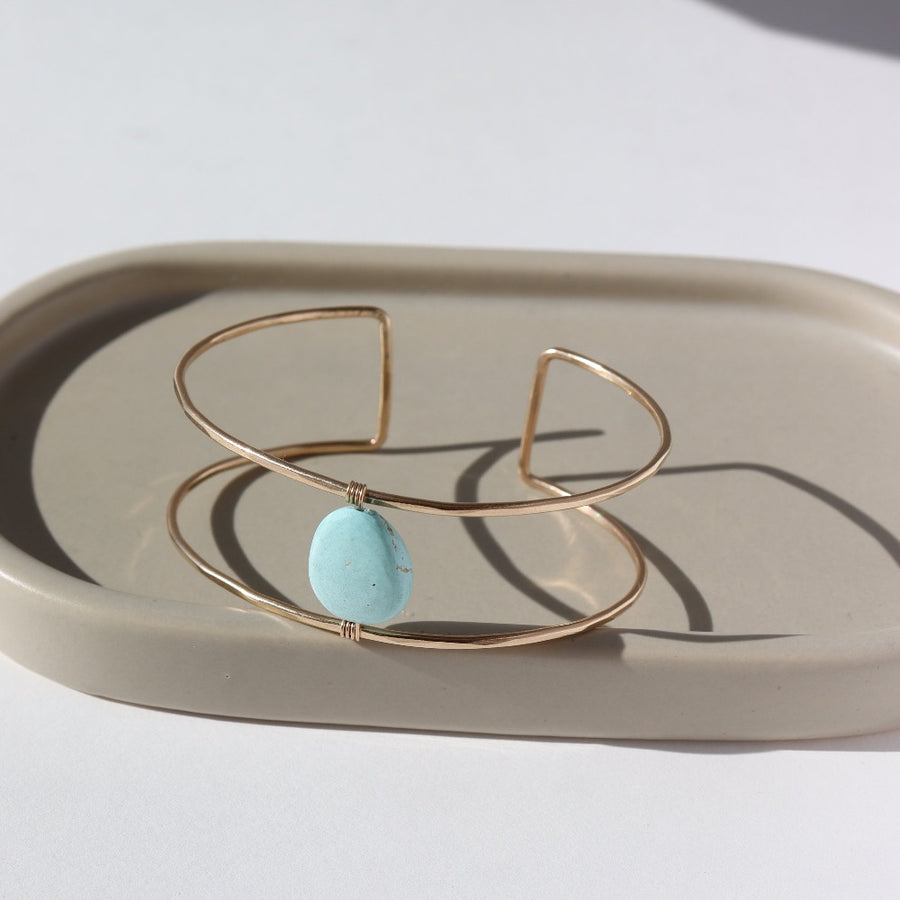 14k gold fill double wire cuff bracelet featuring a large turquoise stone wired to the middle. Photographed on a taupe dish in the sunlight