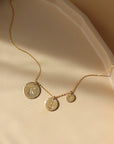 14k gold fill Monogram coin necklace on a peach colored plate in the sunlight.