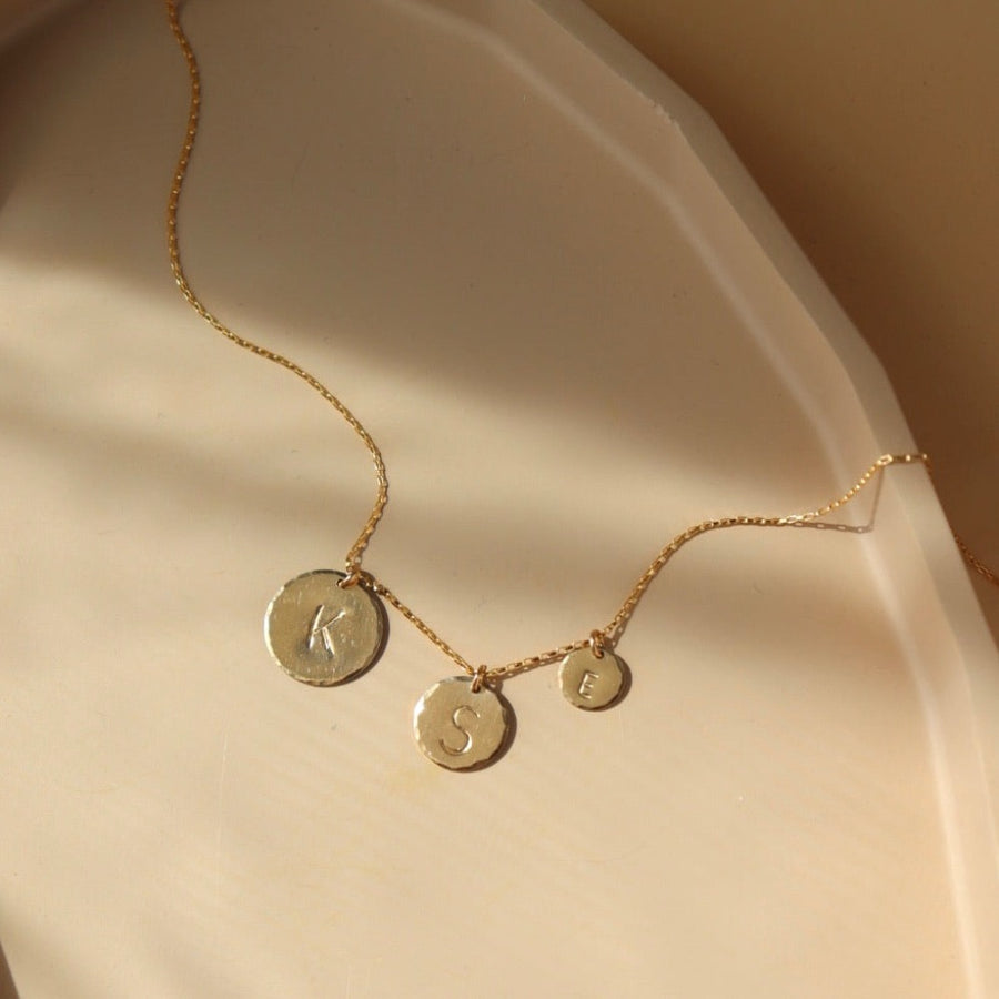 Monogram Coin Necklace laid on a peach colored plate in the sunlight. This necklace shows all different sizes of the Monogram coin. 