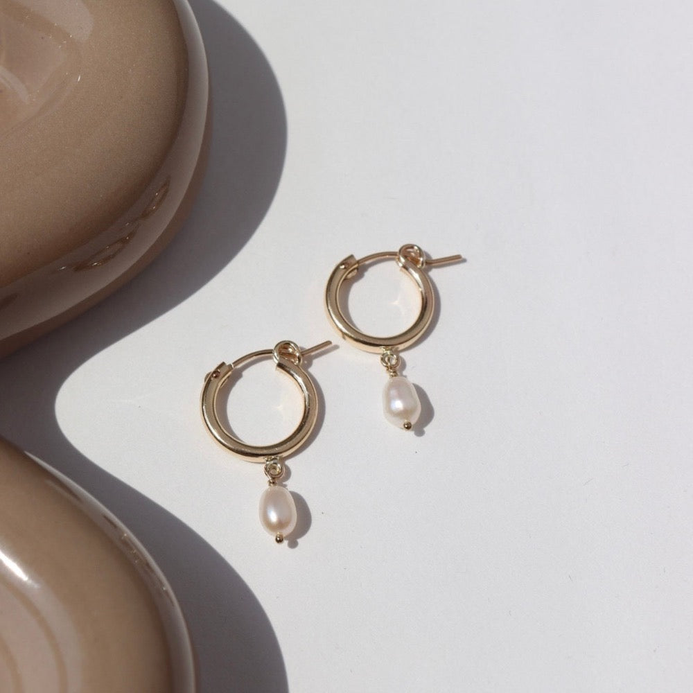 14k gold fill classic pearl hoops laid on a white plate in the sunlight.