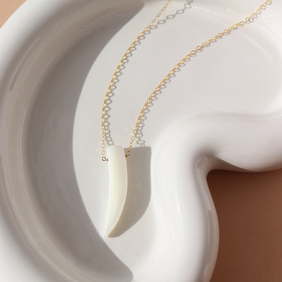 a tusk-shaped pendant made of mother of pearl on a 14k gold fill chain, photographed on a white ceramic dish