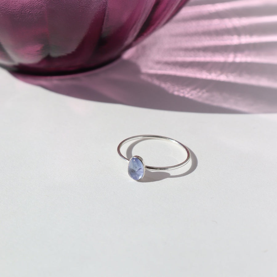 925 Sterling silver Tanzanite Ring laid on a white paper. There is a purple pot in the background of the photo. This Ring features a Smooth band with a Tanzanite Gemstone.
