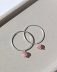 925 Sterling Silver Cherry Blossom Hoops laid on a tan plate in the sunlight. These earring feature a simple hoop earring with the perfect mini gemstone dangle. The gemstone featured in this earring is a Cherry Agate Sphere.