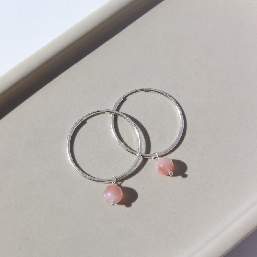 925 Sterling Silver Cherry Blossom Hoops laid on a tan plate in the sunlight. These earring feature a simple hoop earring with the perfect mini gemstone dangle. The gemstone featured in this earring is a Cherry Agate Sphere.