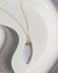14k gold fill Lavender Haze Necklace laid on a white plate in the sunlight. This necklace features the magical Light blue Calcedony stone. This stone is then wrapped in a 14k gold fill wire and dangles off of a simple chain. 