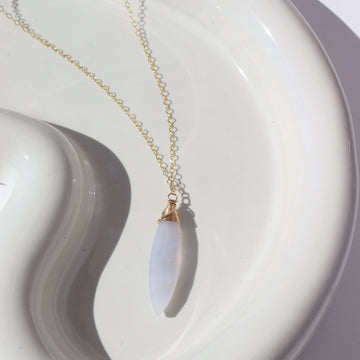 14k gold fill Lavender Haze Necklace laid on a white plate in the sunlight. This necklace features the magical Light blue Calcedony stone. This stone is then wrapped in a 14k gold fill wire and dangles off of a simple chain. 