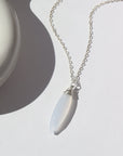 925 Sterling Silver Lavender Haze Necklace laid on a white paper in the sunlight. This necklace features the magical Light blue Calcedony stone. This stone is then wrapped in a 925 Sterling silver wire and dangles off of a simple chain.