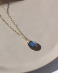 14k gold fill Stormi Necklace laid on a tan plate in the sunlight. This necklace features the simple chain with the Labradorite teardrop gemstone.