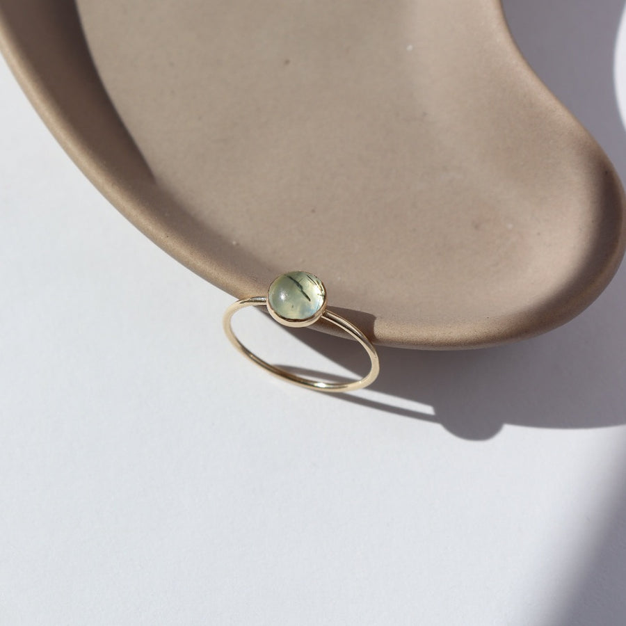 14k gold fill Sweet Pea ring laid on a tan plate in the sunlight. This Ring features a smooth band with a prehnite gemstone.