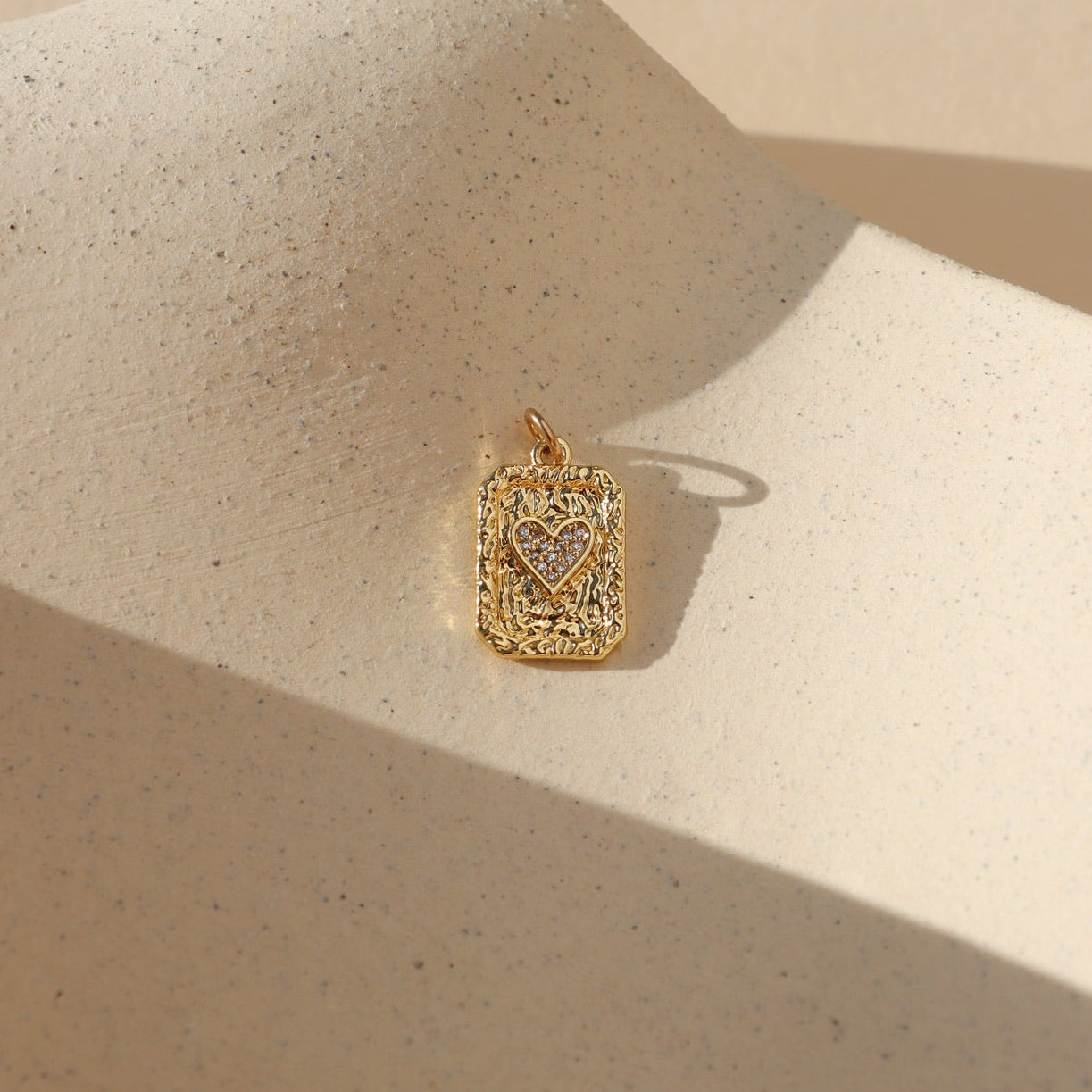 Gold rectangular playing card charm with a textured finish and pavé heart center, photographed on a neutral stone surface in warm natural light.