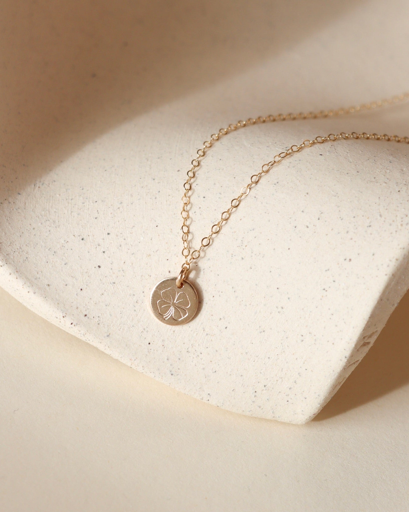 Gold filled necklace with a round hand stamped four leaf clover pendant on a beige stone surface