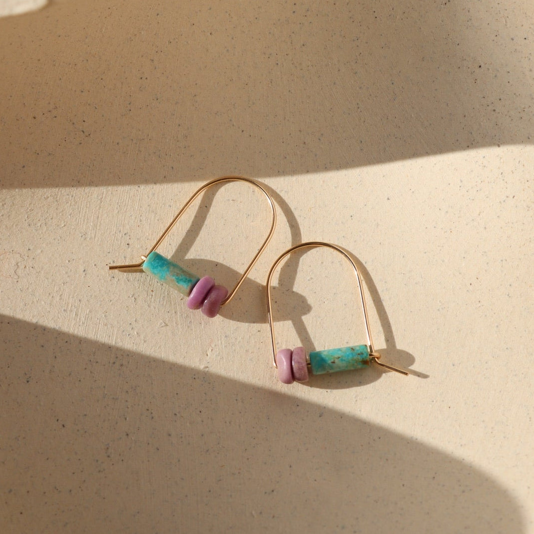 Gold arch-shaped threader earrings featuring turquoise tube beads and phosphosiderite gemstone accents, photographed on a warm cream stone surface in natural sunlight.