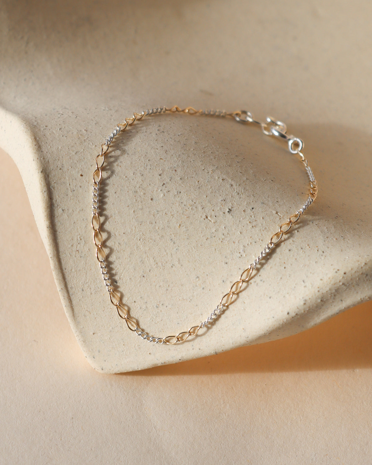 Token Jewelry Interlude Chain Bracelet featuring alternating gold and silver links in a delicate mixed metal design, finished with a lobster clasp and displayed on a textured cream surface in natural sunlight.