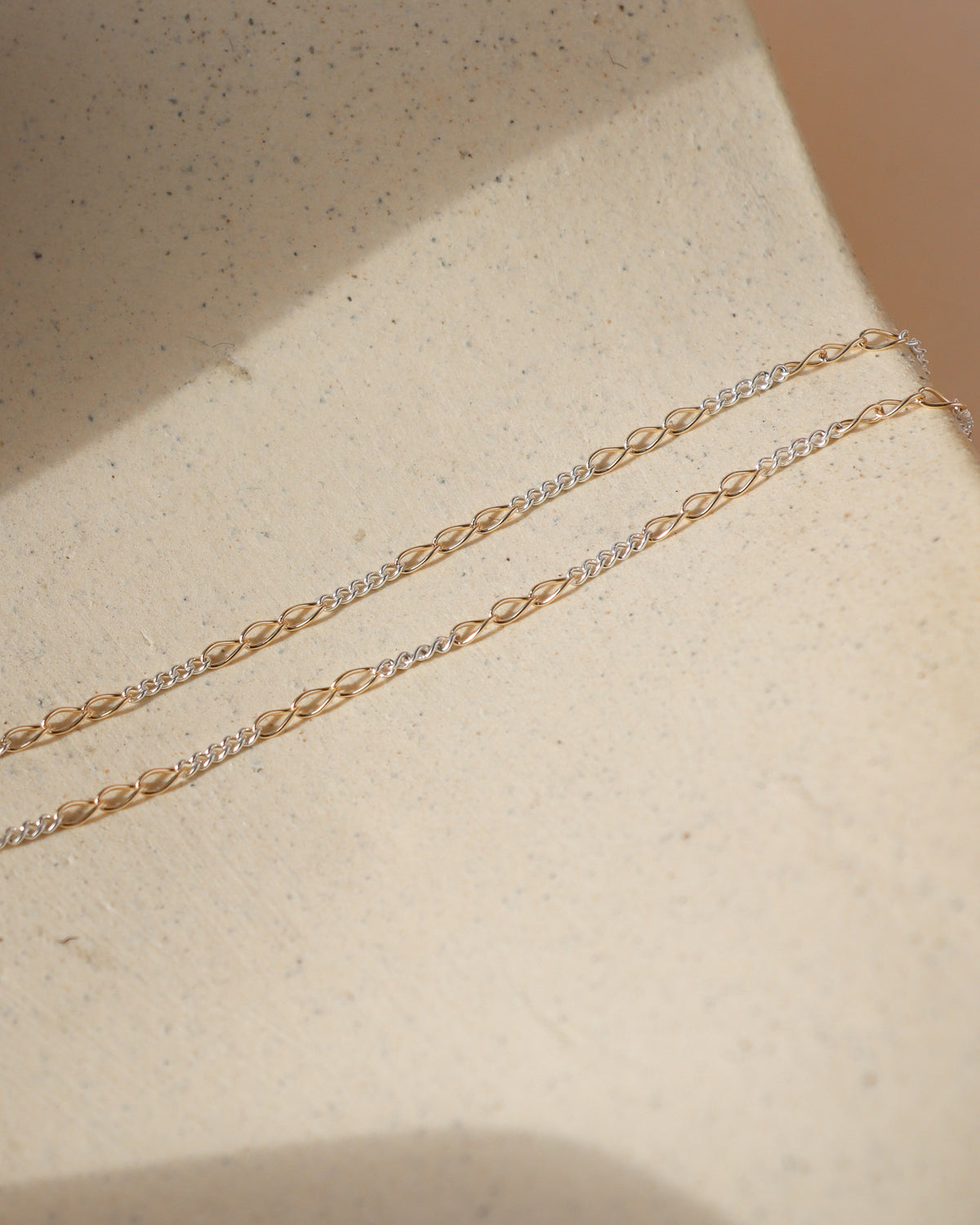 Close-up of Token Jewelry’s delicate mixed-metal chain necklace featuring alternating gold elongated links and silver chain sections, laid across a textured cream stone surface in warm natural sunlight.