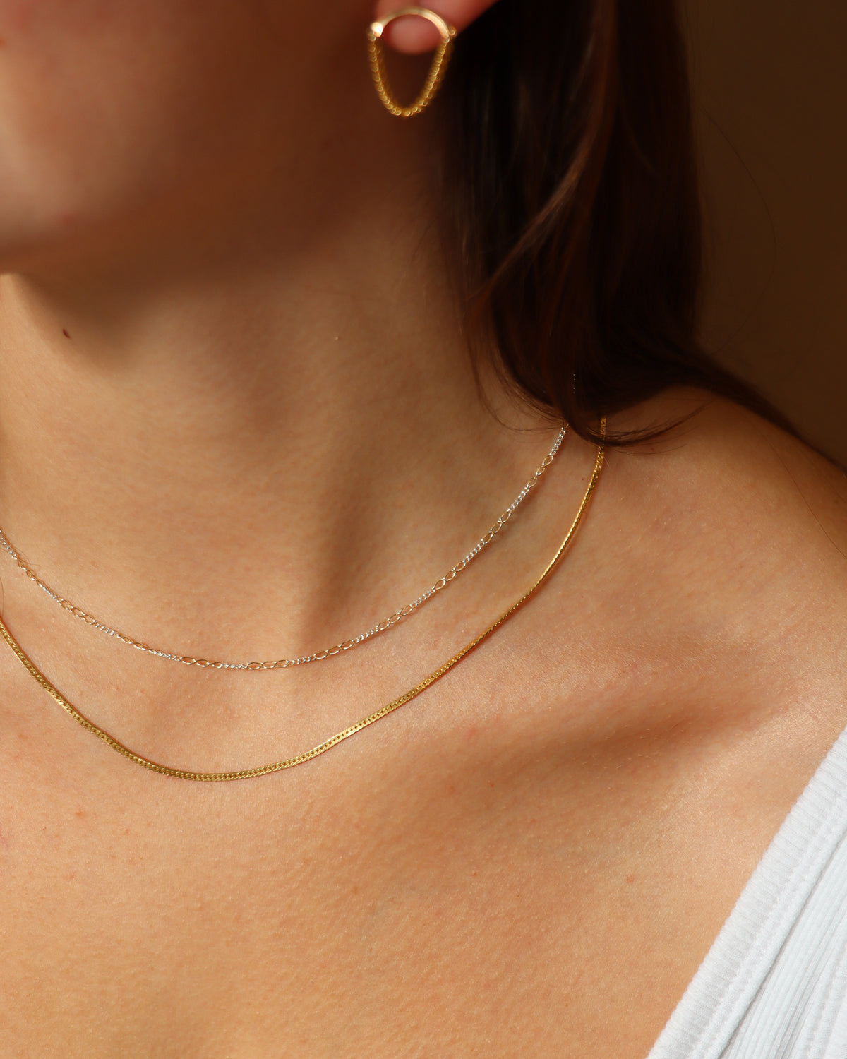 Close-up of a model wearing a delicate mixed-metal chain necklace layered with a gold chain necklace and gold chain drop earrings, styled in warm natural sunlight.