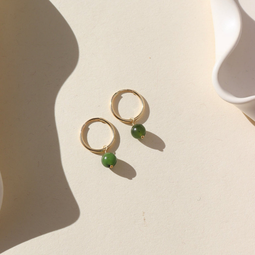 A 14k gold fill 14mm hoop with a Jade ball gemstone dangling from it. Earrings are displayed on a cream background. 