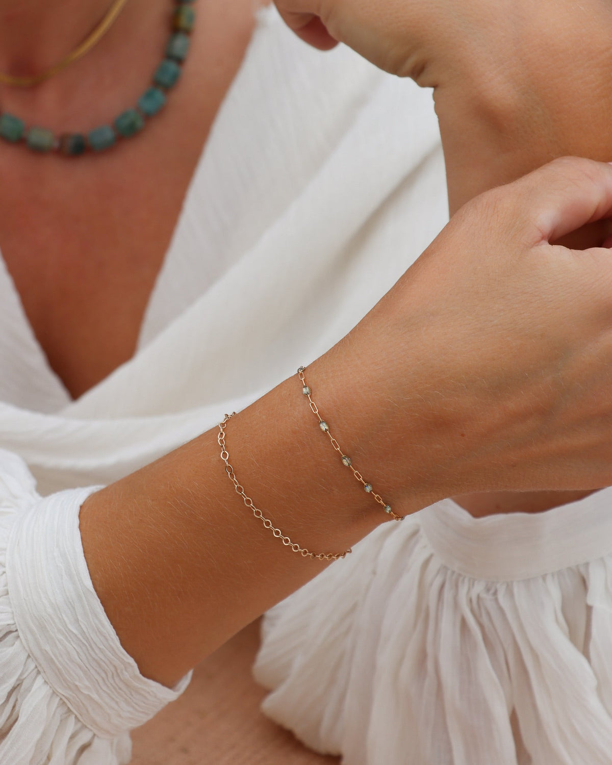 Close-up of layered delicate gold bracelets styled with a green beaded necklace and white blouse.