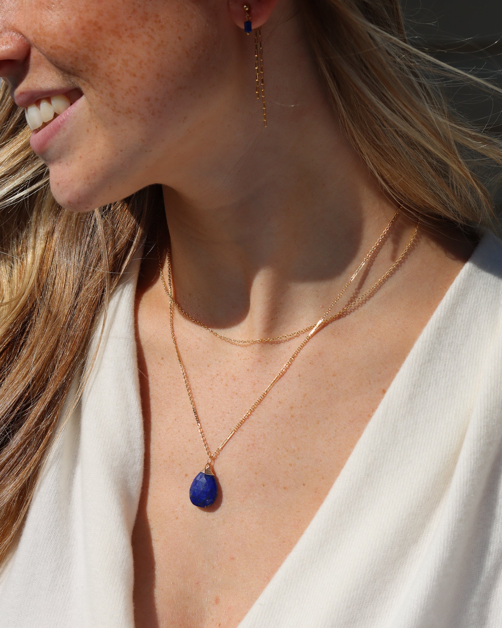 A woman wearing a layered gold necklace featuring a teardrop-shaped lapis lazuli pendant, paired with matching gold drop earrings and a white top.
