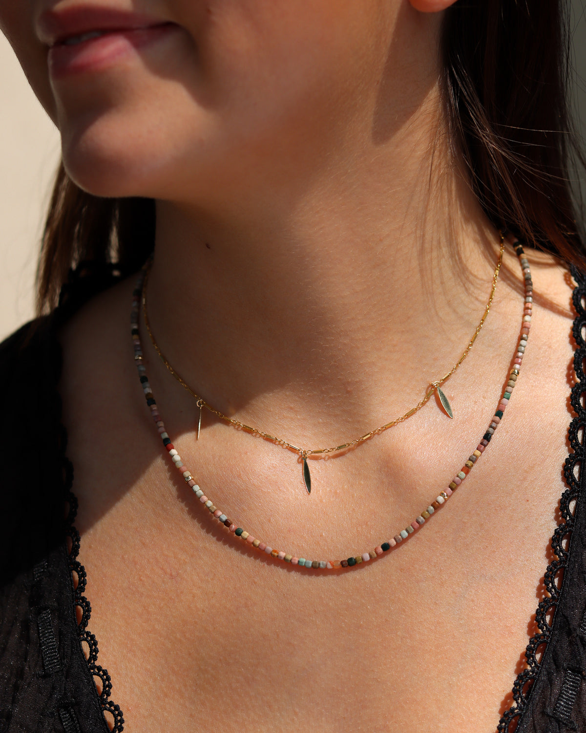 Model wearing a multicolored gemstone beaded necklace layered beneath a delicate gold charm necklace and styled with a black top.